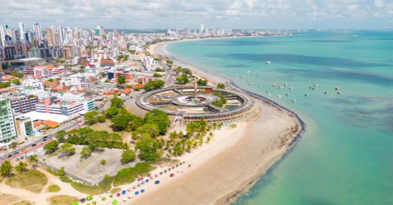 Vista aérea da praia de Tambaú com o icônico Hotel Tambaú em formato redondo na areia, mostrando o mar verde e a orla de João Pessoa. Imagem ilustrativa para o artigo sobre o Polo Turístico em João Pessoa.