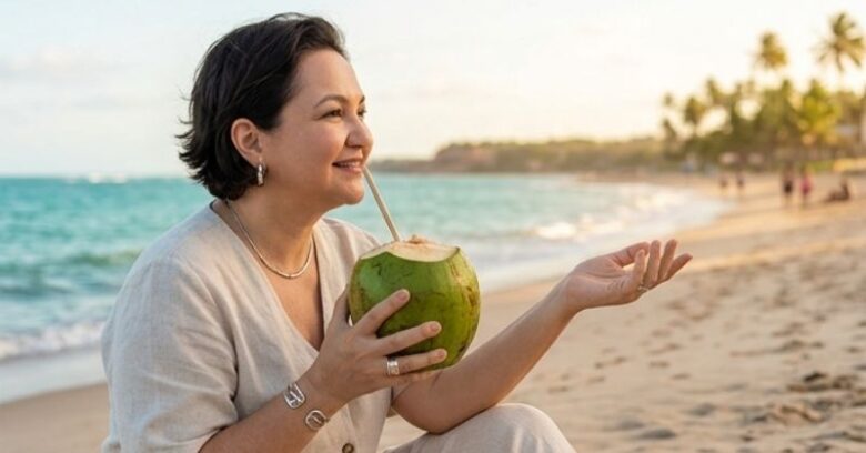 Uma mulher (Graça Pontes)sorridente, sentada na areia da praia de Cabo Branco em João Pessoa, tomando água de coco fresca diretamente da fruta enquanto olha para o mar ao pôr do sol, ilustrando o estilo de vida de quem decide morar na Paraíba.